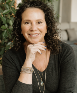 Brunette with curly hair in a grey shirt resting a hand under her smiling mouth.